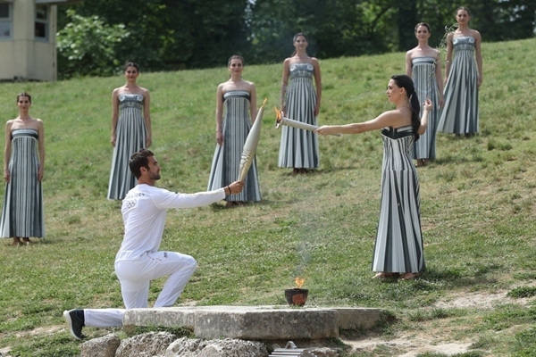 扮演最高女祭司的演員瑪麗·米娜（前右）點燃第一棒火炬手——希臘運動員、東京奧運會男子賽艇項目金牌得主斯特凡諾斯·恩圖斯科斯手中的火炬。新華社記者 趙丁喆 攝